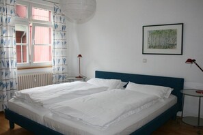 A double bed with white linens, a blue headboard, a window with pink curtains, a small bedside table with a red lamp, and a framed painting on the wall. - Holiday apartment with sea view, Villa Vineta 14 (Seebad Bansin)