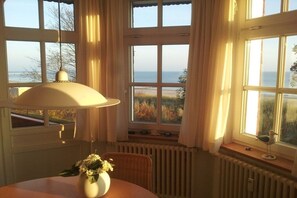 A room with a table, a lamp, a vase with flowers, a wicker chair, and a view of the sea through large windows. - Holiday apartment with sea view, Villa Vineta 14 (Seebad Bansin)
