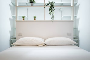 Ein schlicht eingerichtetes Schlafzimmer mit einem Bett, zwei Nachttischen, einem Fenster mit Regalen und einer hängenden Pflanze.