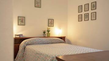 A bedroom with a floral bedspread, a bedside table with a lamp, and framed botanical prints on the wall.
