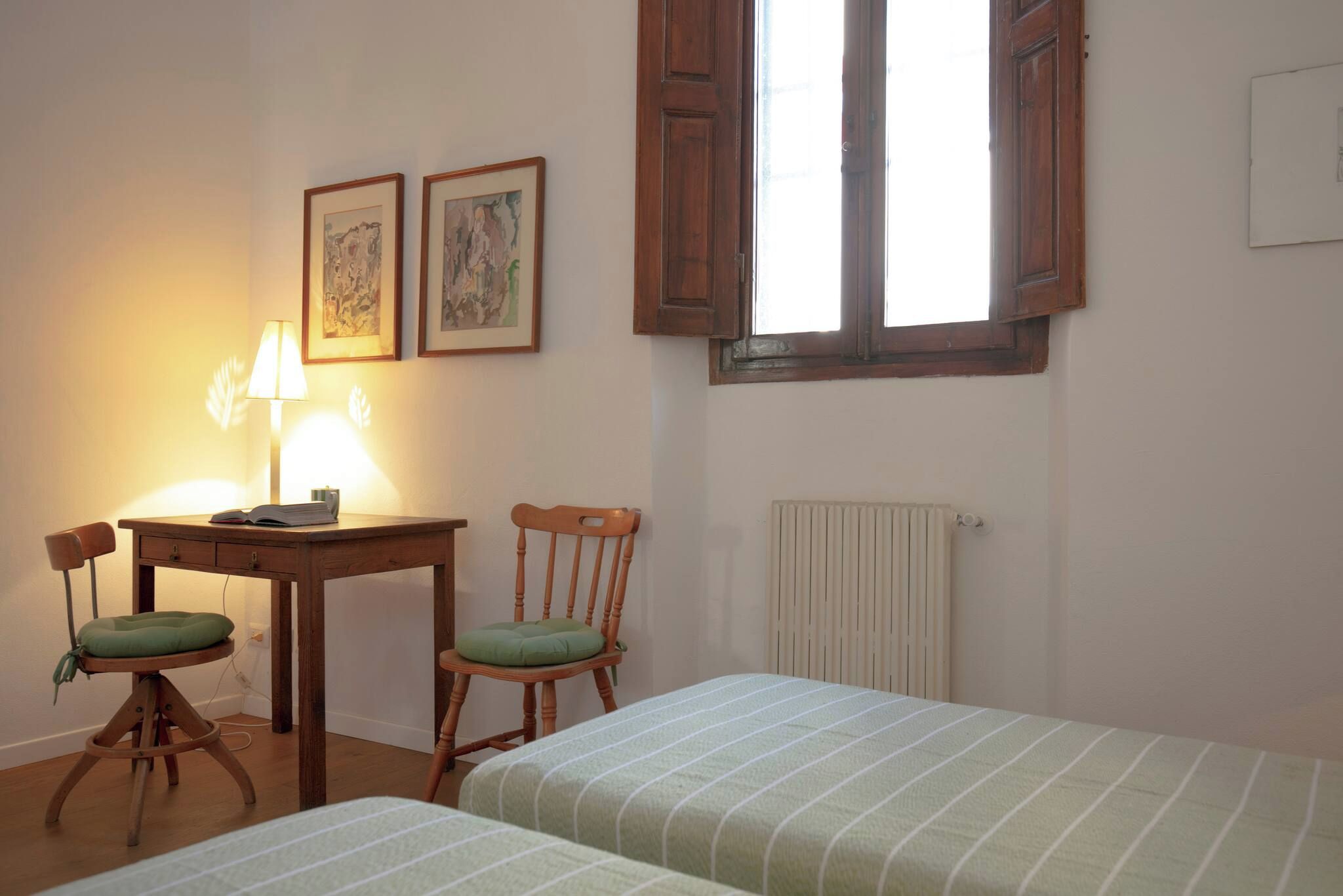 A room with a wooden desk, two chairs, a radiator, and two beds.