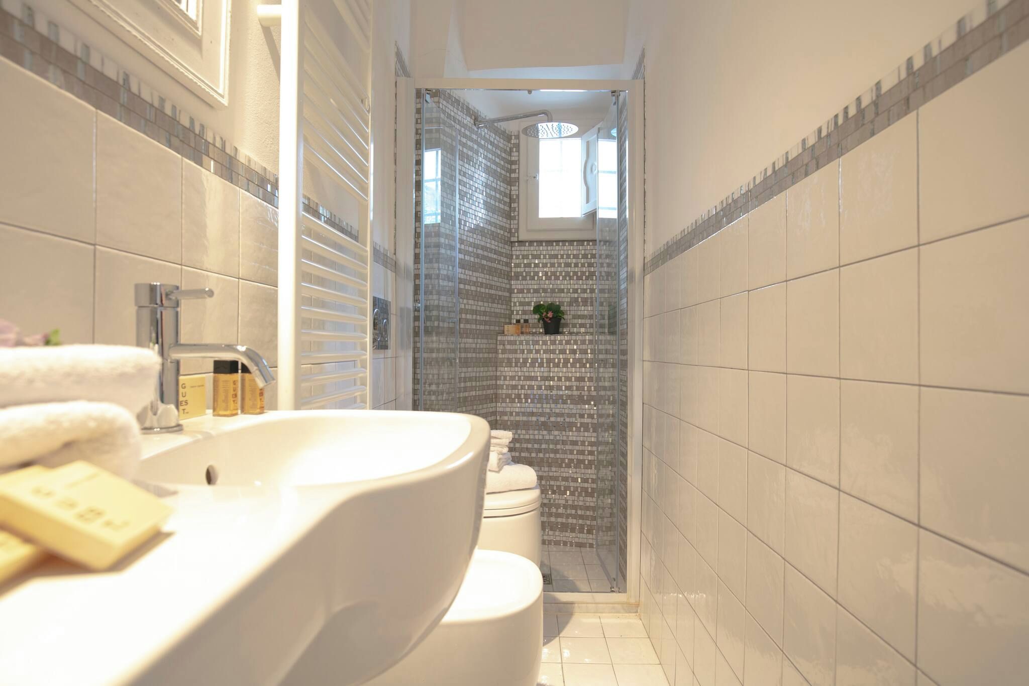 Ein schmales Badezimmer mit einem weißen Waschbecken, einer Toilette und einer Duschkabine mit Glastür.