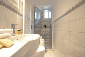 Un baño estrecho con un lavamanos blanco, un inodoro y una zona de ducha con puerta de cristal.