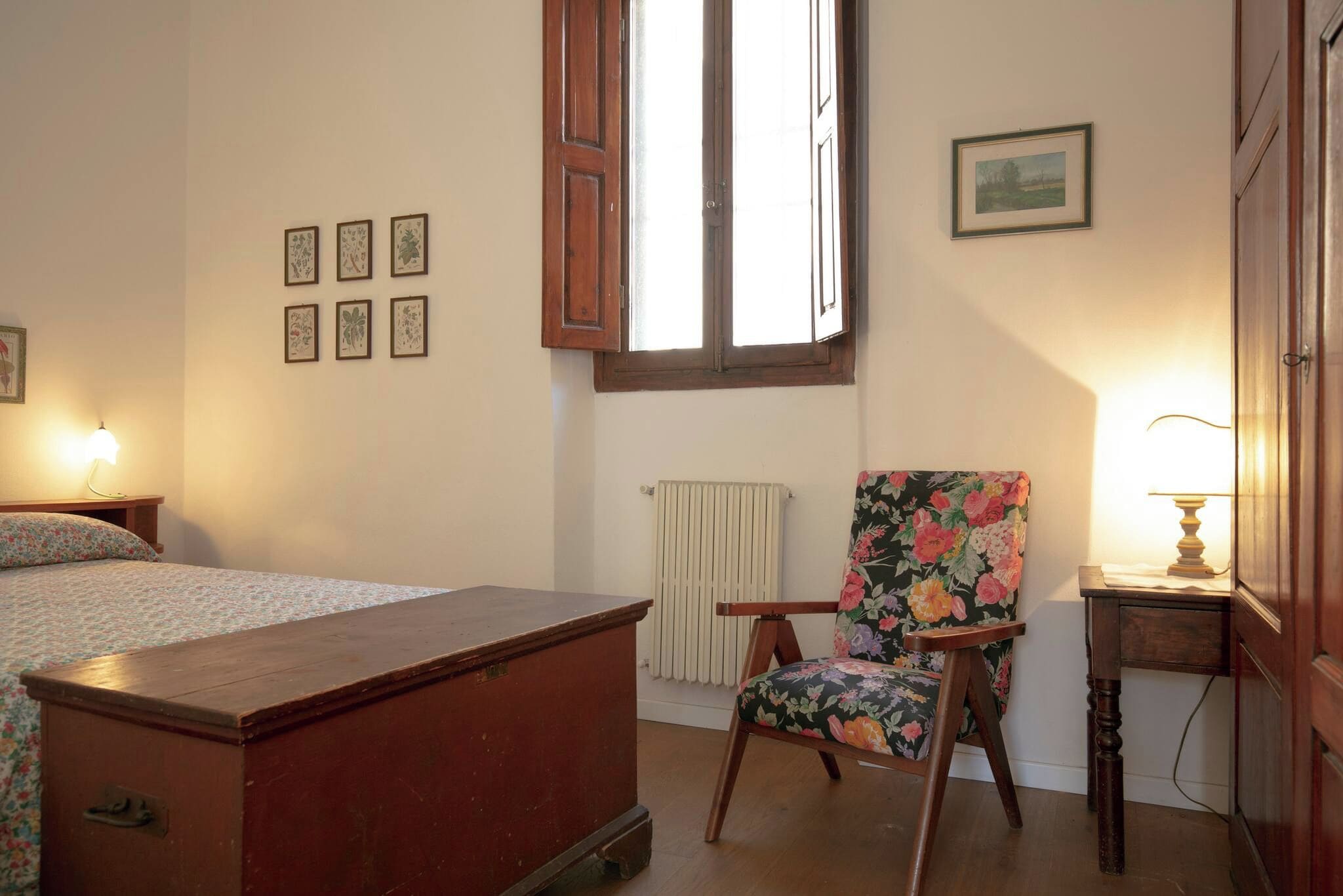 A bedroom with a wooden bed, a floral armchair, a radiator, and framed pictures on the wall.