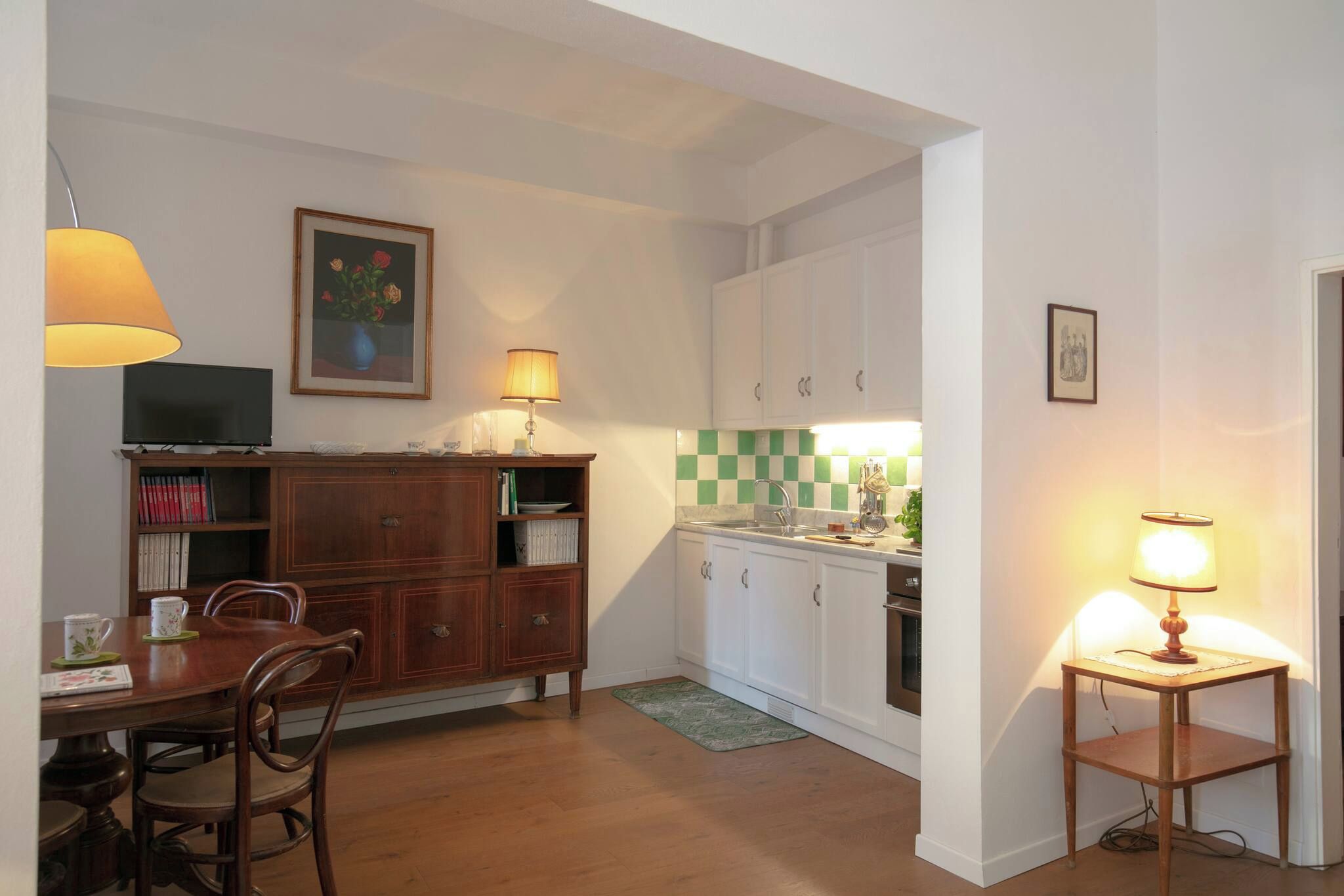 Cocina compacta con armarios blancos, una mesa de comedor de madera y una mesita auxiliar con lámpara.