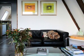 A living room with a black leather sofa, a glass coffee table, and a bookshelf with books. There are two framed pictures on the wall, and a plant in a vase on the floor. - Holiday home with 1 bedroom (Hollern-Twielenfleth)