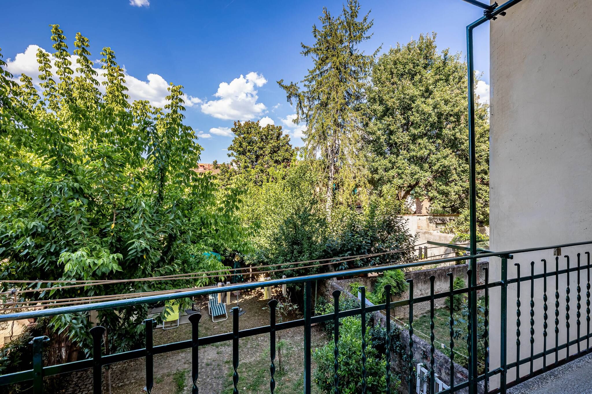 A balcony with a view of a garden, a stone wall, and a metal railing.