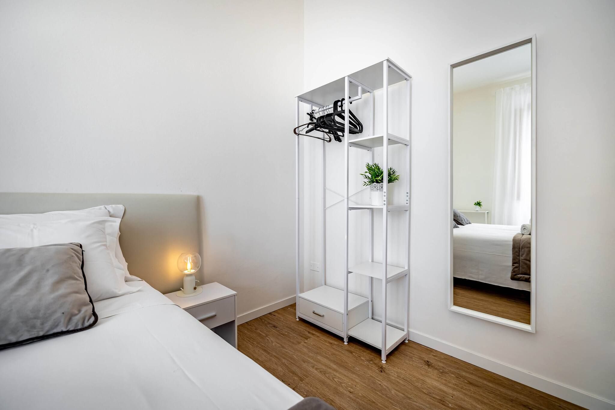 A bedroom with a bed, bedside table, nightstand, white wardrobe, and a full-length mirror.