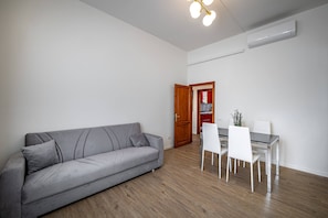 A modern living room with a grey sofa, white dining table, and chairs. A wooden door leads to a kitchen area. - Aleardi (Florence)