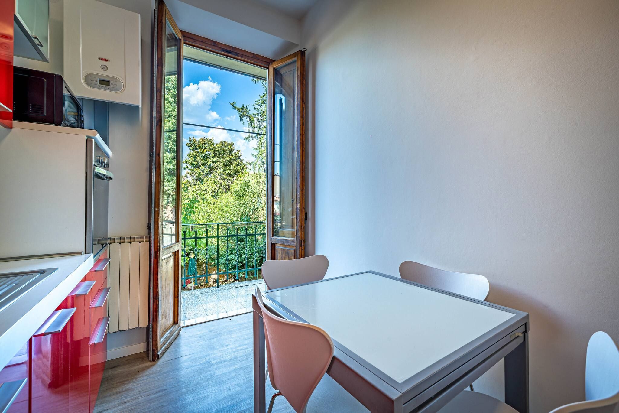 A modern kitchen with a built-in microwave, a white refrigerator, and a red appliance on the counter. There is a glass table with two chairs and a view of a balcony with greenery outside.