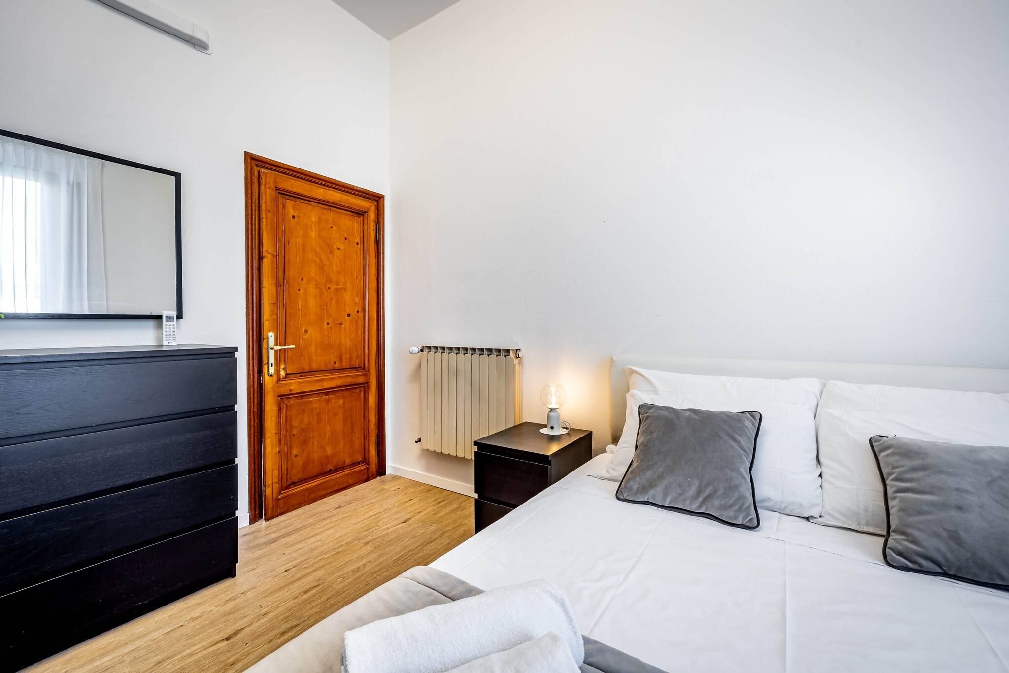 A bedroom with a wooden wardrobe, a bedside table with a lamp, a bed with white and grey bedding, and a radiator.