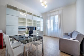 A modern living room with a glass-top dining table, white chairs, a grey sofa, and built-in shelving. - Aleardi (Florence)