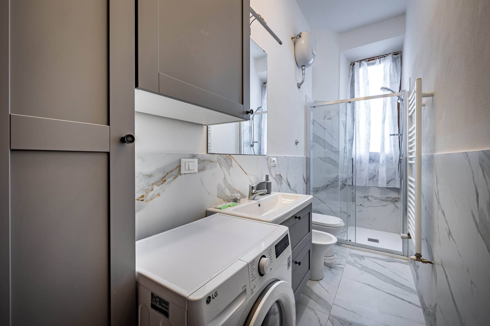 Une salle de bain moderne dotée d’un lave-linge intégré, d’une douche en verre et d’un sol carrelé en marbre.