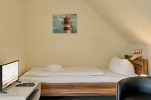 Uma cama de solteiro com cabeceira de madeira, uma televisão sobre um suporte, um urso de pelúcia em uma mesa de cabeceira e uma pintura de um farol.