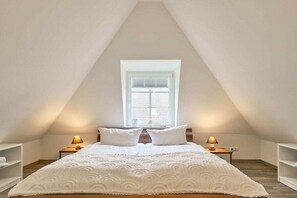 Ein Schlafzimmer mit schrägen Wänden, einem Bett mit weißer Bettwäsche, zwei Nachttischlampen und einem Fenster mit Jalousien.