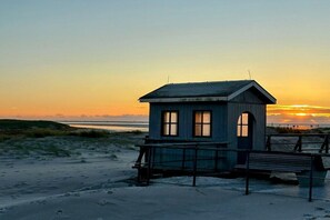 Eine kleine Holzkonstruktion mit geneigtem Dach, einer Bank und einem Geländer, gelegen an einem Sandstrand mit Sonnenuntergang im Hintergrund.
