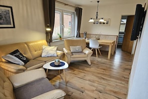 A living room with a sofa, armchair, and a small round table. There is a dining area with a table and chairs. A window with curtains is visible, and a TV is mounted on the wall. - North Sea holiday Stormhus (Sankt Peter-Ording)