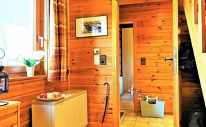 Ein Holzhütten-Innenraum mit einer Holztür, einem kleinen Tisch mit Obst, einem an der Wand befestigten Lichtschalter und einem Bilderrahmen.