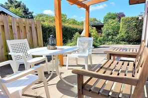 Eine Terrasse im Freien mit weißen Kunststoffstühlen, einer Holzbank, einem kleinen Tisch und einer Laterne.