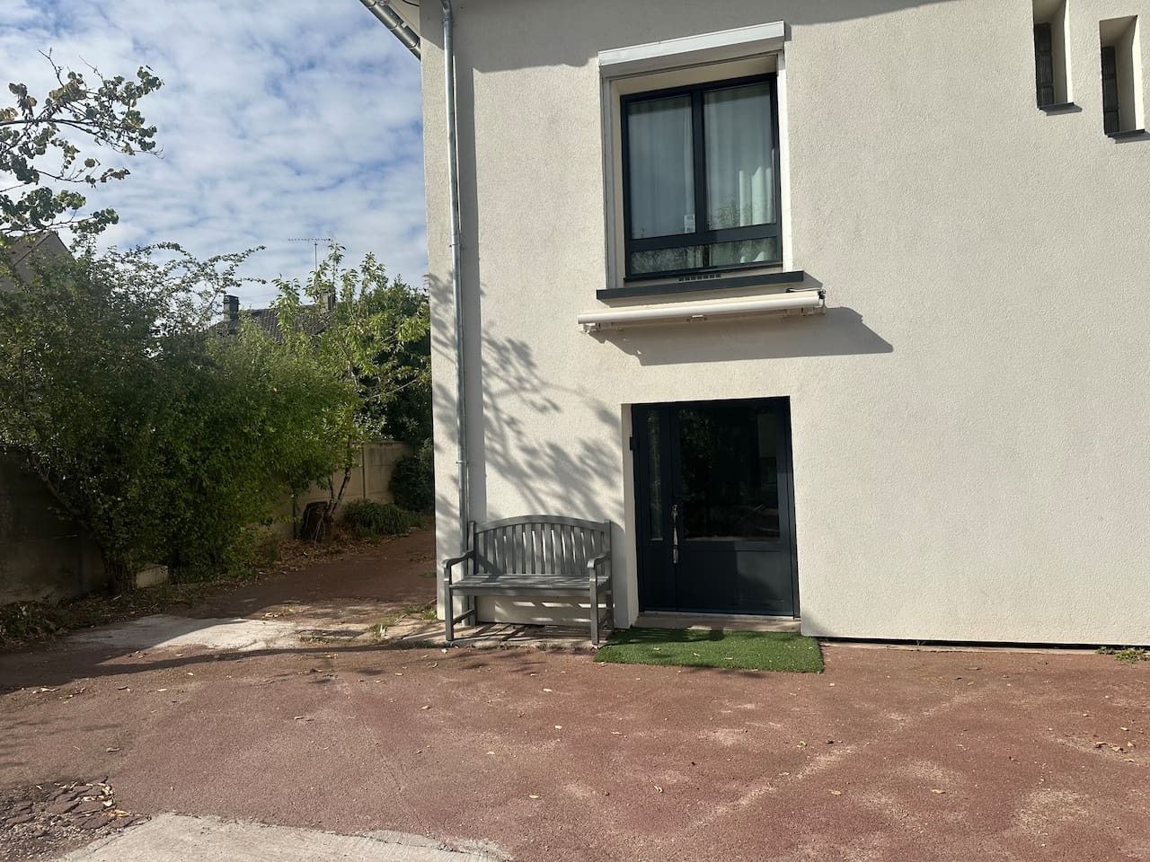 Een gebouw met één raam en een deur, een bankje ervoor en een verhard gebied.