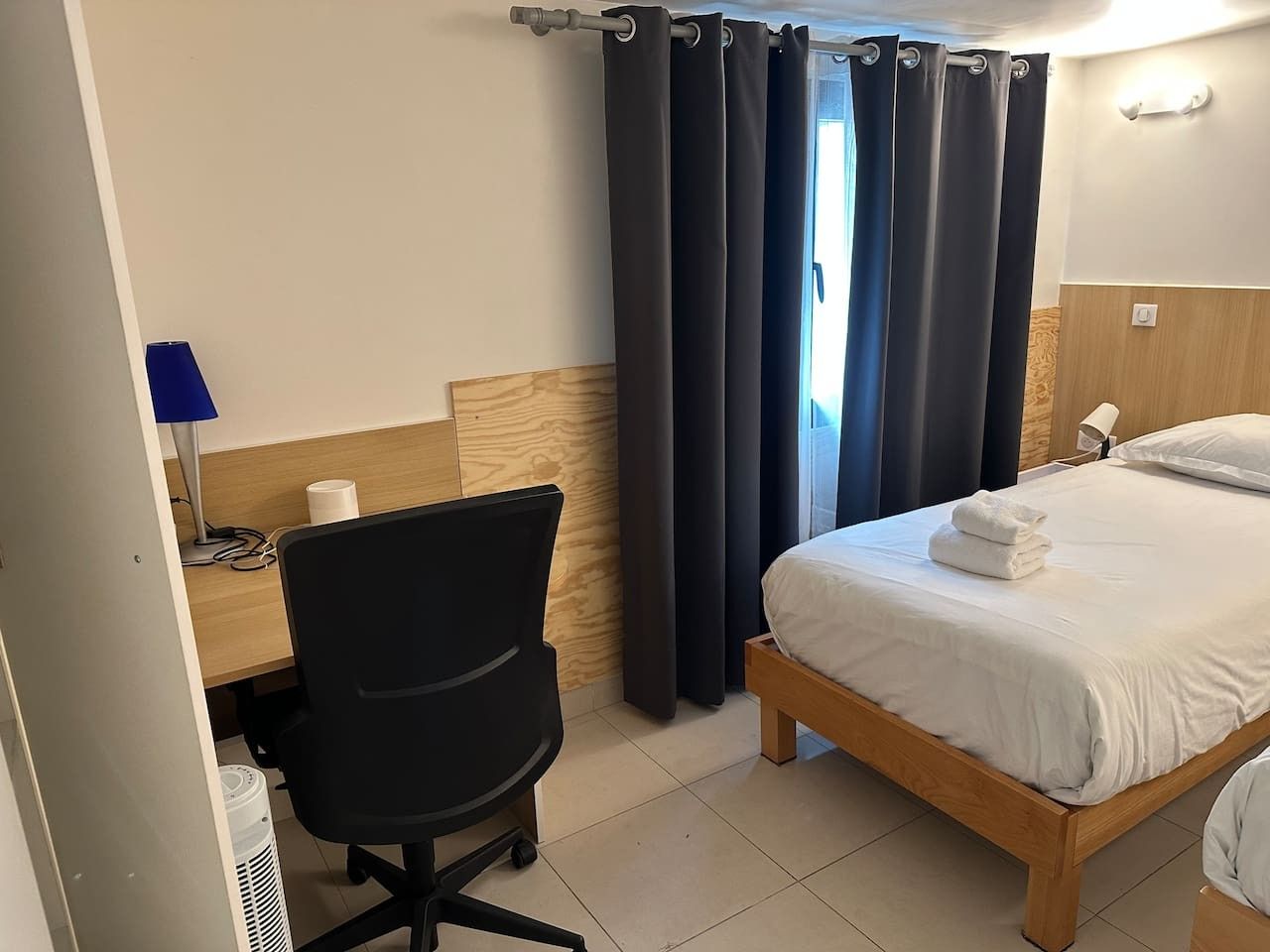 Una camera d'albergo con un letto, una scrivania con una sedia, una lampada e una finestra con tende.