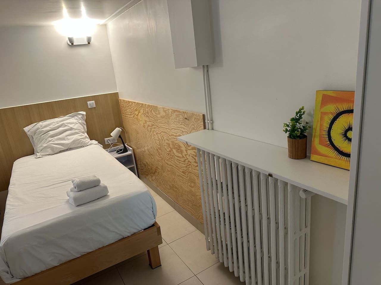 Una camera d'albergo con un letto, un comodino, una lampada, un termosifone e un'opera d'arte appesa al muro.