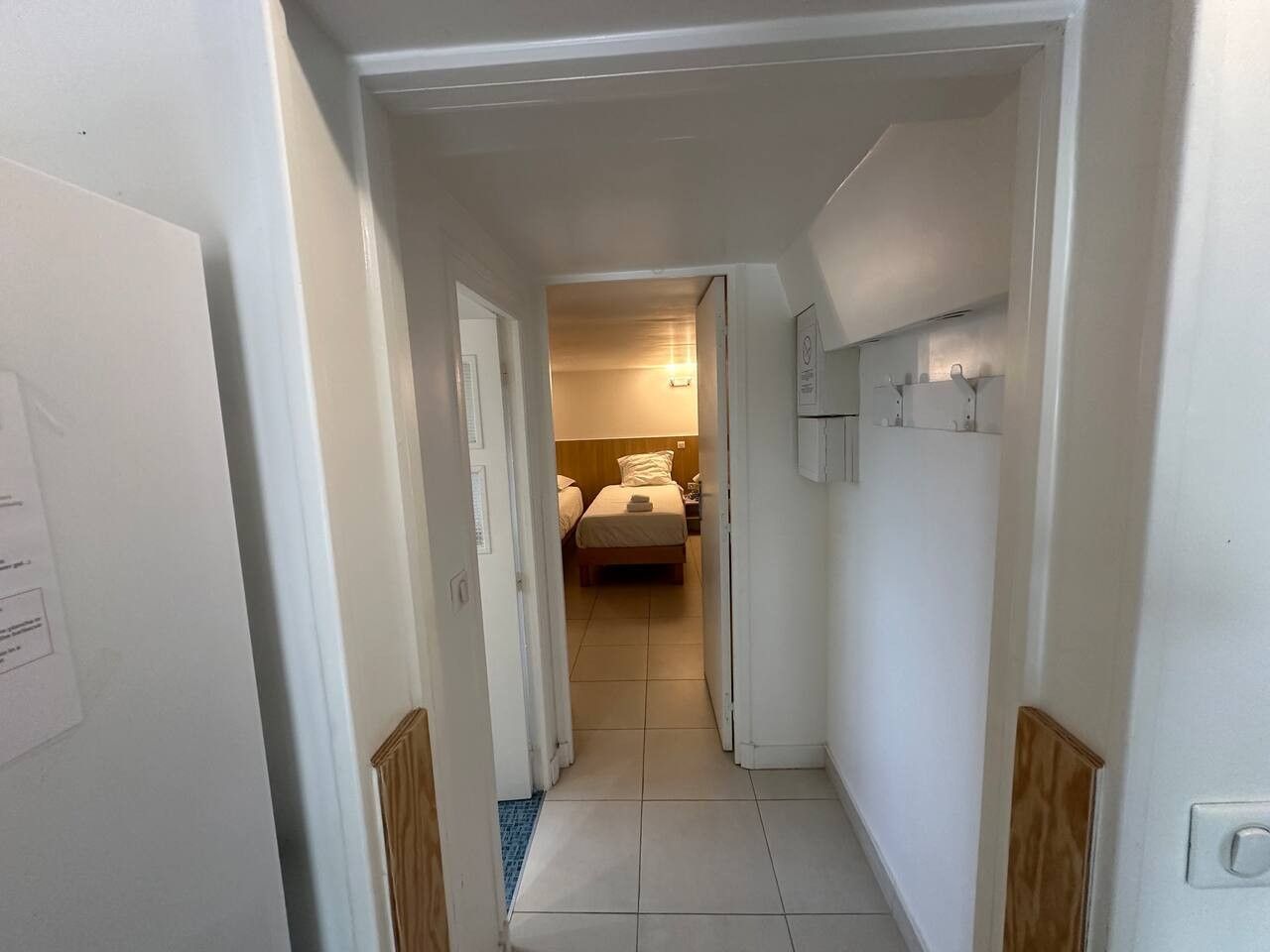 Een smalle gang die leidt naar een slaapkamer met een bed, een nachtkastje en een wandlamp.