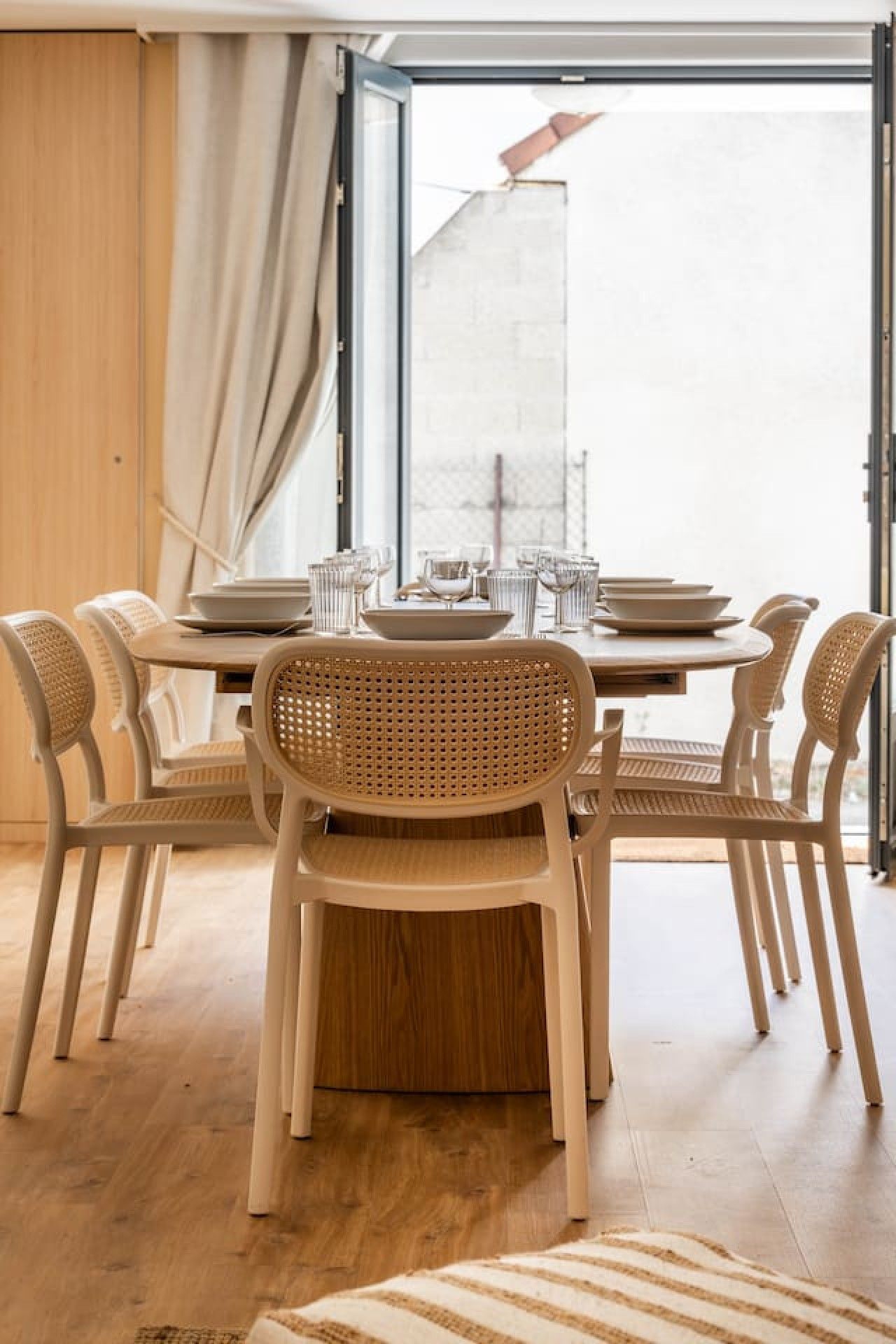 A dining area with a table set for four, surrounded by wicker chairs, and a view through a glass door.