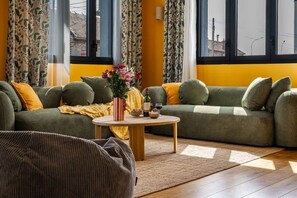 A cozy living room with a green sofa, yellow cushions, a striped vase with flowers, and a small table with a bottle and glasses. - Spacious Apartment—5 Bedrooms—Accommodates 10 Guests—20 Minutes from Paris—2 Parking Spaces (Argenteuil)
