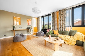 A modern living room with a sofa, coffee table, and bean bag chairs. There is a desk with a laptop and a vase with flowers. The room has large windows with curtains. - Spacious Apartment—5 Bedrooms—Accommodates 10 Guests—20 Minutes from Paris—2 Parking Spaces (Argenteuil)
