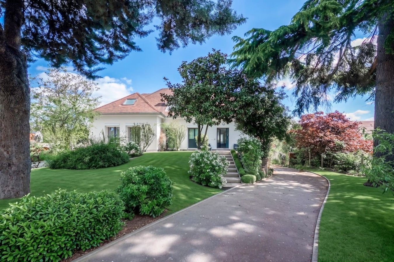 A two-story house with a well-maintained lawn, a curved driveway, and a variety of trees and shrubs.