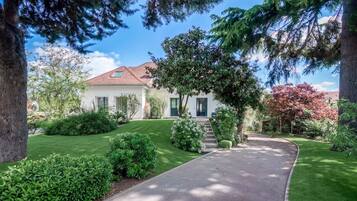 A two-story house with a well-maintained lawn, a curved driveway, and a variety of trees and shrubs.