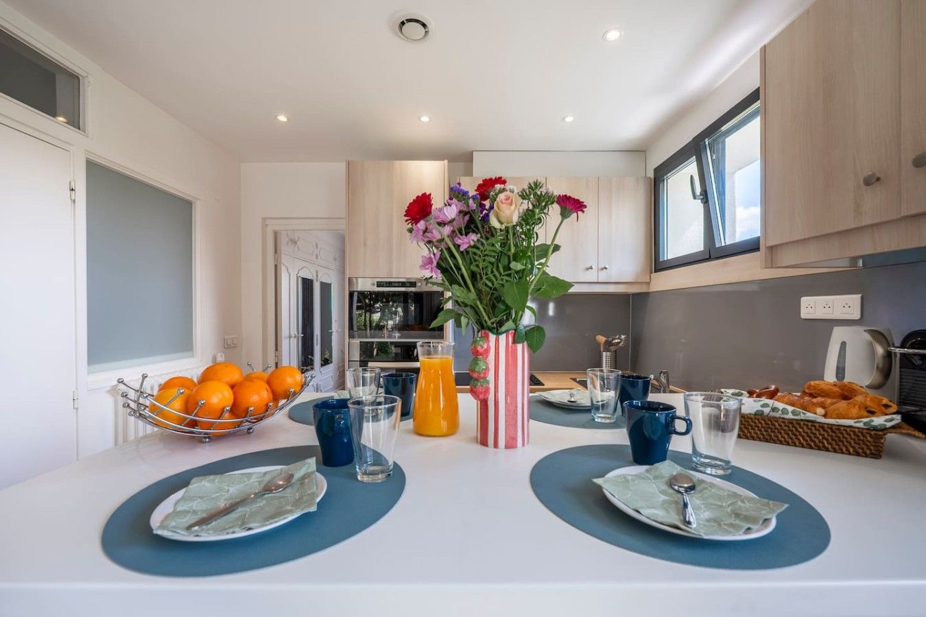A modern kitchen with a dining table set for two, featuring a fruit bowl, a vase with flowers, and a basket of pastries.