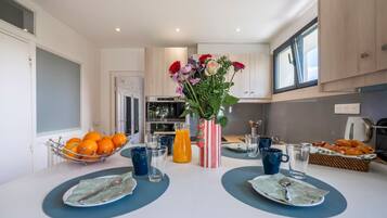 A modern kitchen with a dining table set for two, featuring a fruit bowl, a vase with flowers, and a basket of pastries.