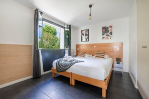 A modern bedroom with a large bed, a window with curtains, a bedside table, and artwork on the wall.