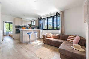 Eine moderne Küche mit einer zentralen Kochinsel, einem Essbereich mit einem braunen Sofa und einem großen Fenster, das natürliches Licht hereinlässt.