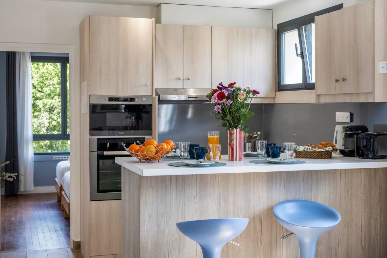 Eine moderne Küche mit Holzschränken, einer zentralen Kochinsel mit Barhocker und einem Essbereich mit Frühstücksbereitstellung, darunter Obst, eine Blumenvase und ein Toaster.