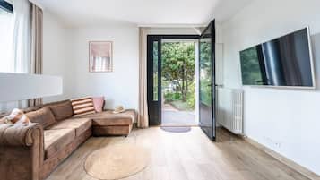 Un salon moderne avec un canapé d’angle, un téléviseur à écran plat et une vue sur un jardin, accessible par une porte ouverte.