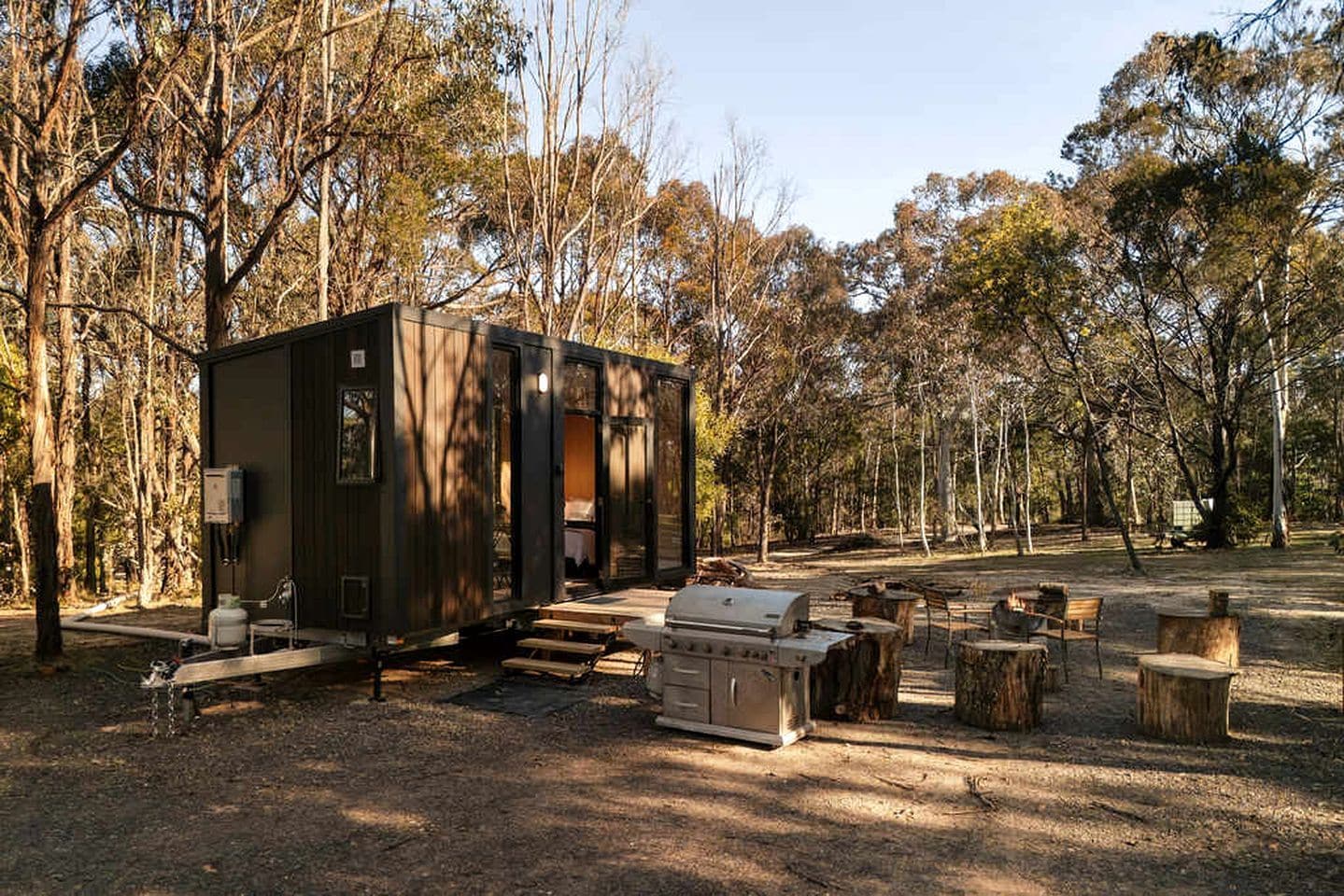 A small cabin with a grill and outdoor seating, surrounded by trees.