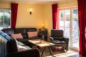 A living room with a sofa set, a coffee table, and a chair. There is a window with curtains, a wall-mounted light, and a small table with a lamp.