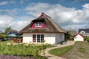 A thatched-roof house with a red window and white walls, surrounded by a garden and other houses.
