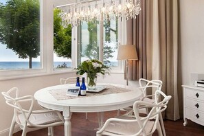 A dining area with a round table, white chairs, a chandelier, a vase with flowers, and a view of the sea through large windows. - Beach jewel - Villa Agnes (Binz)