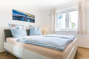 Una cama con ropa de cama a rayas, una mesita de noche, una lámpara y un cuadro de un puente en la pared.