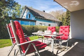 Un patio con una mesa y dos sillas rojas, una casa azul al fondo y un seto en primer plano.
