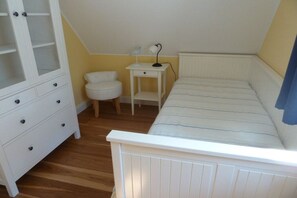 Ein Schlafzimmer mit einem weißen Bett, einem Nachttisch mit Lampe, einer weißen Kommode und einem kleinen, runden Stuhl.