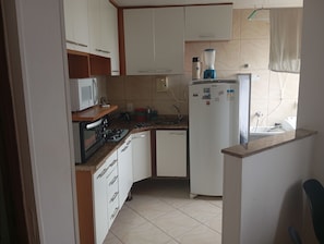 A compact kitchen with white cabinets, a microwave, a stove, a refrigerator, and a blender. - Very quiet and safe apartment near Barra, 24-hour doorman, shops. (Rio de Janeiro)