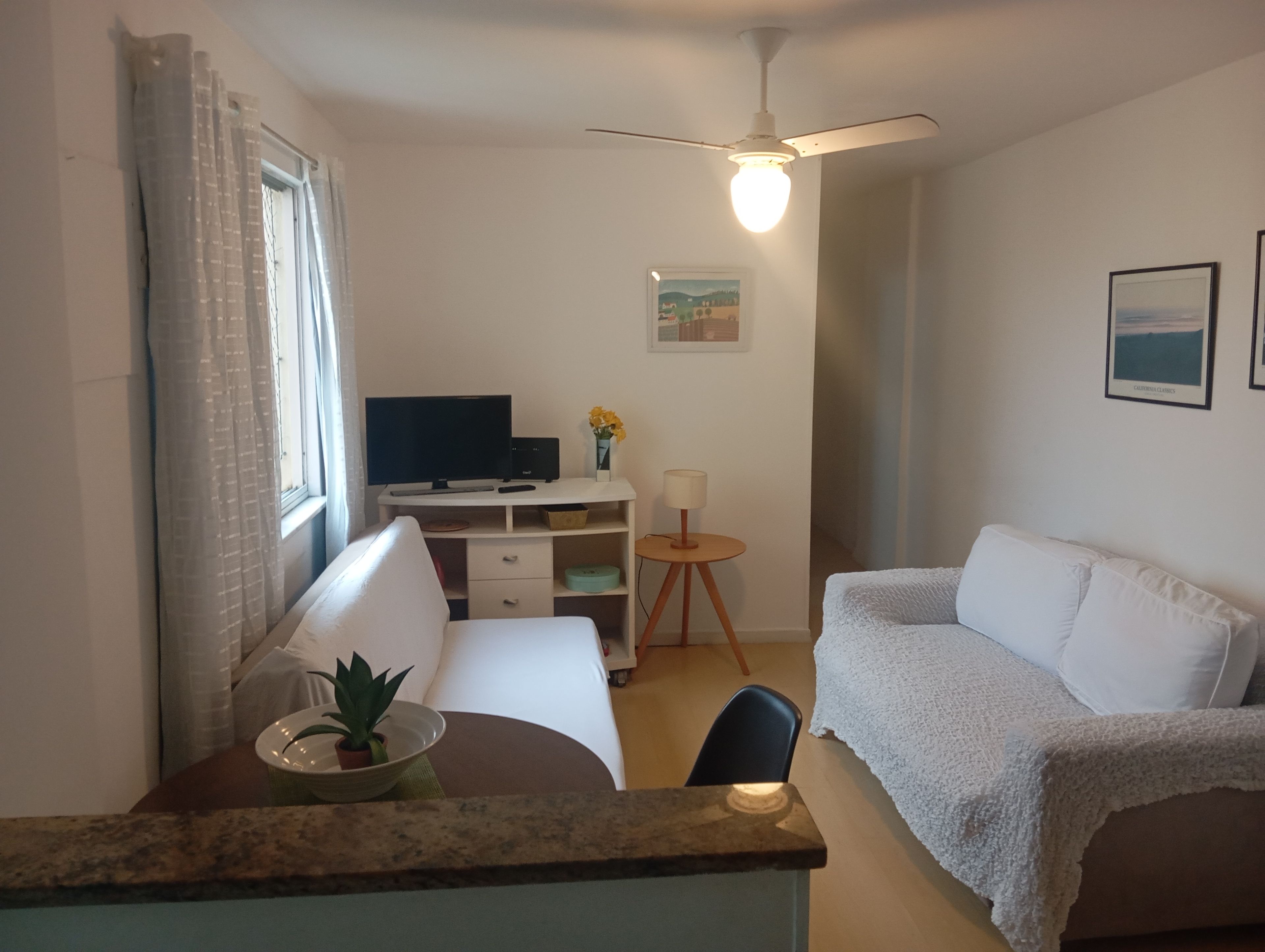 A room with a bed, a desk with a computer, a chair, a small table with a lamp, and a ceiling fan.