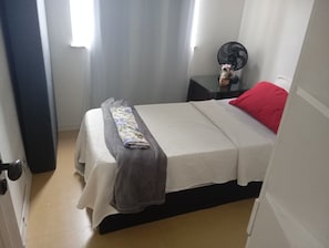 Ein kleines, ordentlich eingerichtetes Schlafzimmer mit einem Einzelbett, einem Nachttisch mit Ventilator und einem roten Kopfkissen.