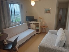 A room with a white sofa, a TV on a stand, a small table with a lamp, and a painting on the wall. - Very quiet and safe apartment near Barra, 24-hour doorman, shops. (Rio de Janeiro)