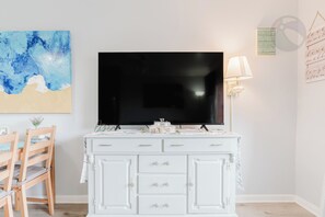 Een wit kastje met lades, een flatscreen televisie erboven bevestigd, een lamp aan de muur en een blauw abstract schilderij.